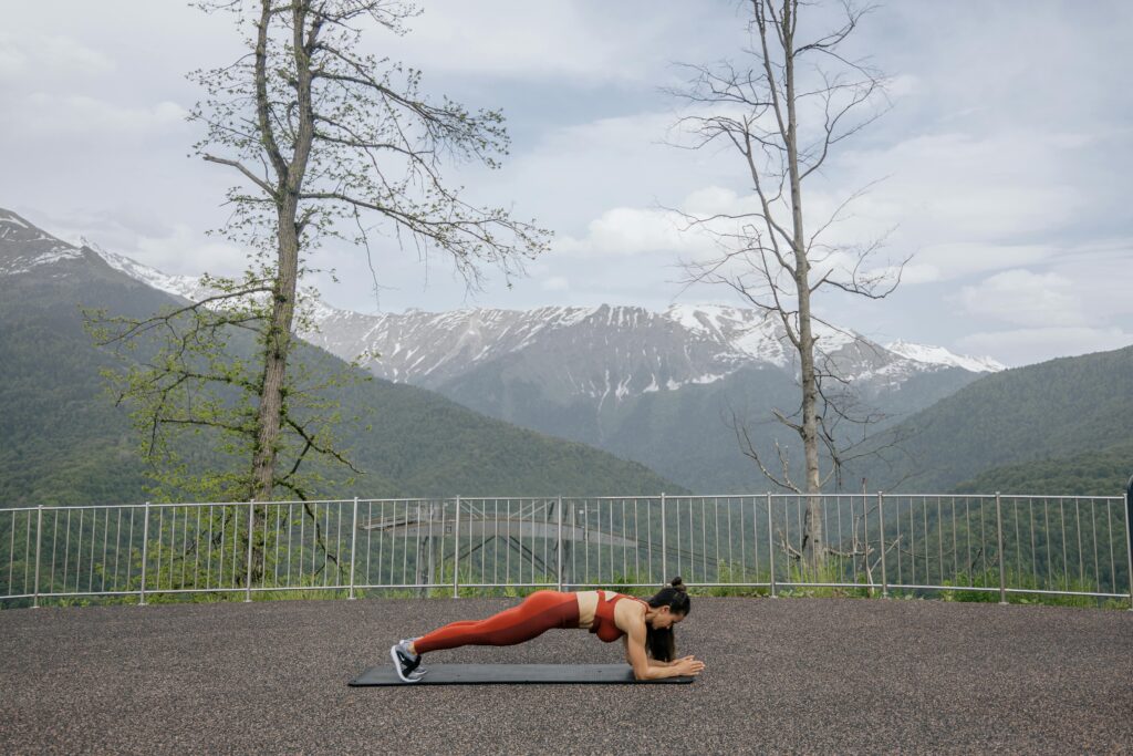 A woman doing a plank exercise on a mat outdoors with a scenic mountain view in the background.